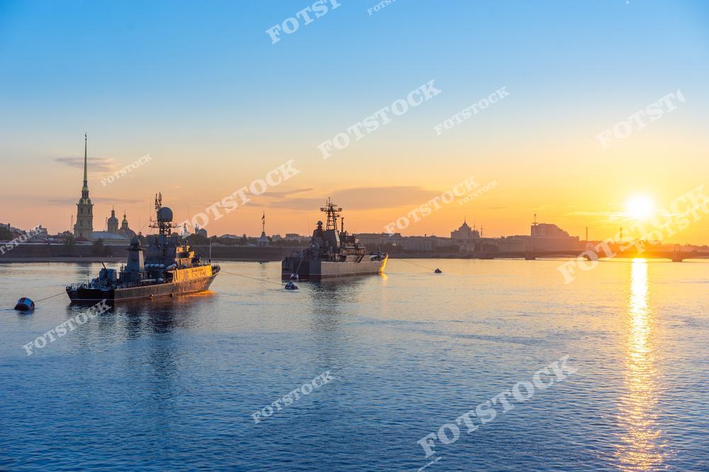 На фотографии изображены военные корабли, стоящие на якоре в водах реки Невы в Санкт-Петербурге. На заднем плане видна Петропавловская крепость с её знаменитым золотым шпилем. Закатное солнце создает теплое свечение, отражающееся на воде и придающее сцене торжественность и спокойствие. Этот вид на Неву символизирует морскую мощь России и красоту вечернего Петербурга.